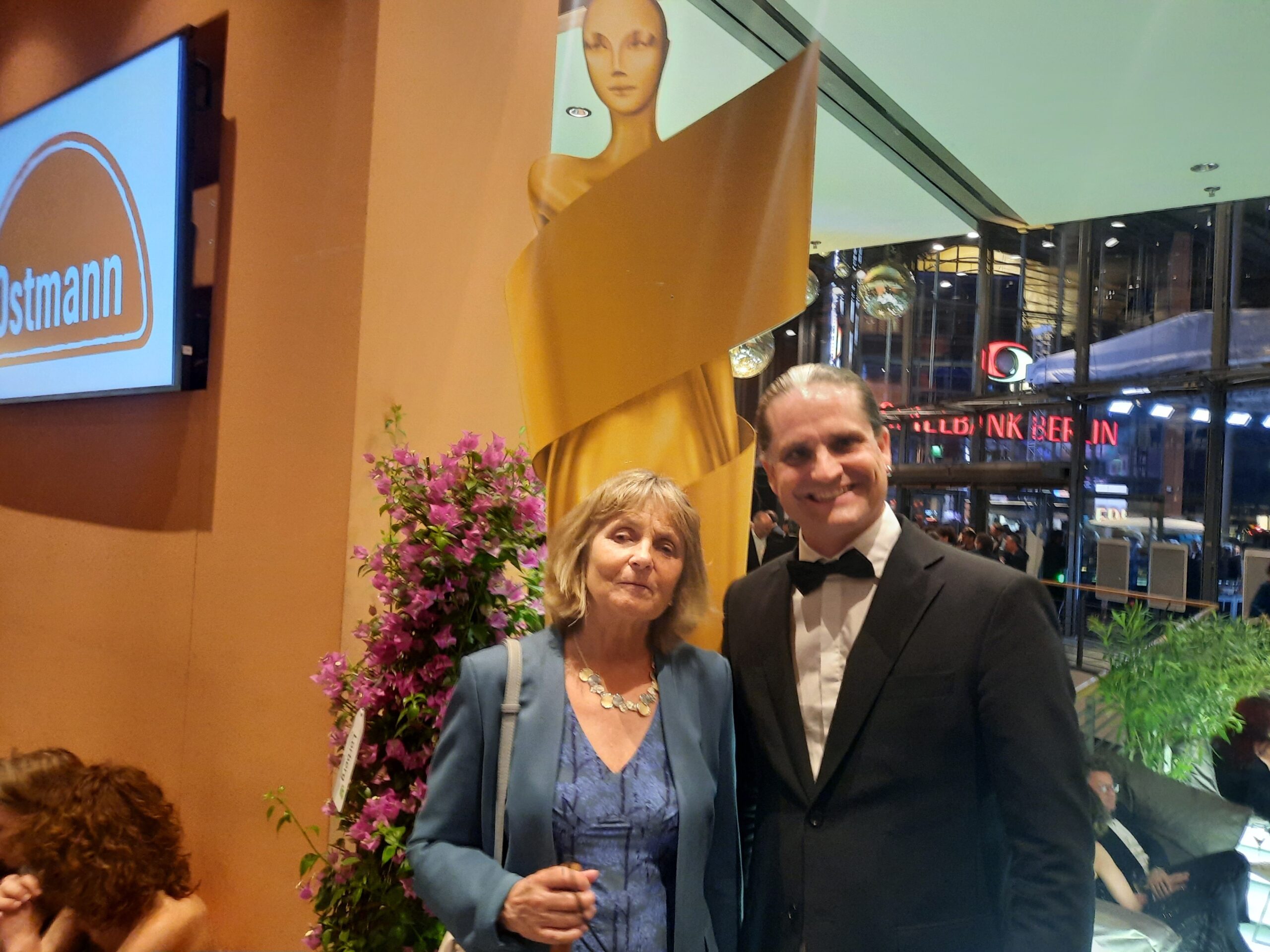 Die Blindgängerin und Andres Schüpbach von der Greta App stehen im Foyer des Theaters am Potsdamer Platz. Sie lächeln in die Kamera. Mittig hinter ihnen eine große goldene Statue der Lola.